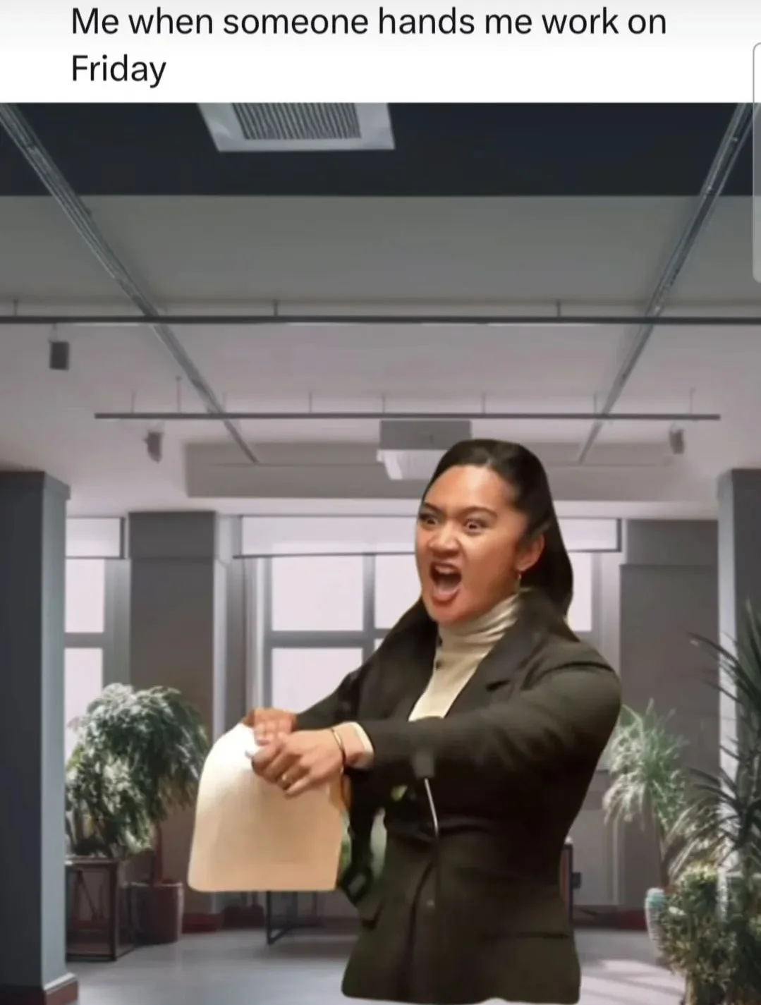 A woman in a black blazer and cream turtleneck stands in an office hallway, holding a beige folder, her face contorted in…