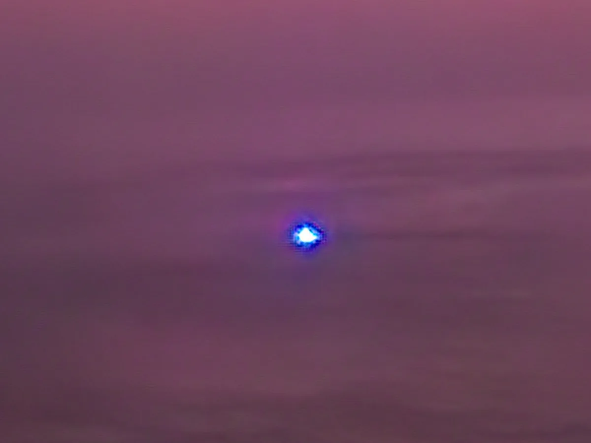 A blurry, low-resolution image shows a single bright blue light piercing through a hazy, purple-pink sky, resembling a…