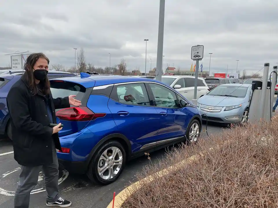 A person wearing a black face mask and a black jacket stands beside a blue electric car in a parking lot, gesturing toward…