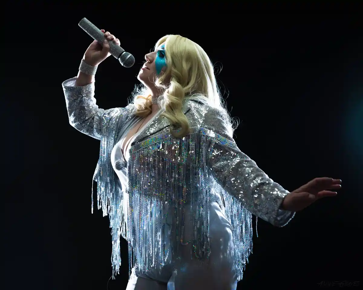 A person with blonde hair and blue face paint wears a sparkly silver fringe jacket and holds a glittery microphone while…