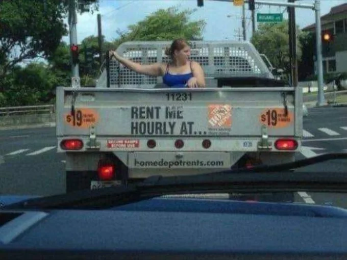 A woman in a blue tank top leans out the back of a silver utility truck stopped at a traffic light. The truck's tailgate has…