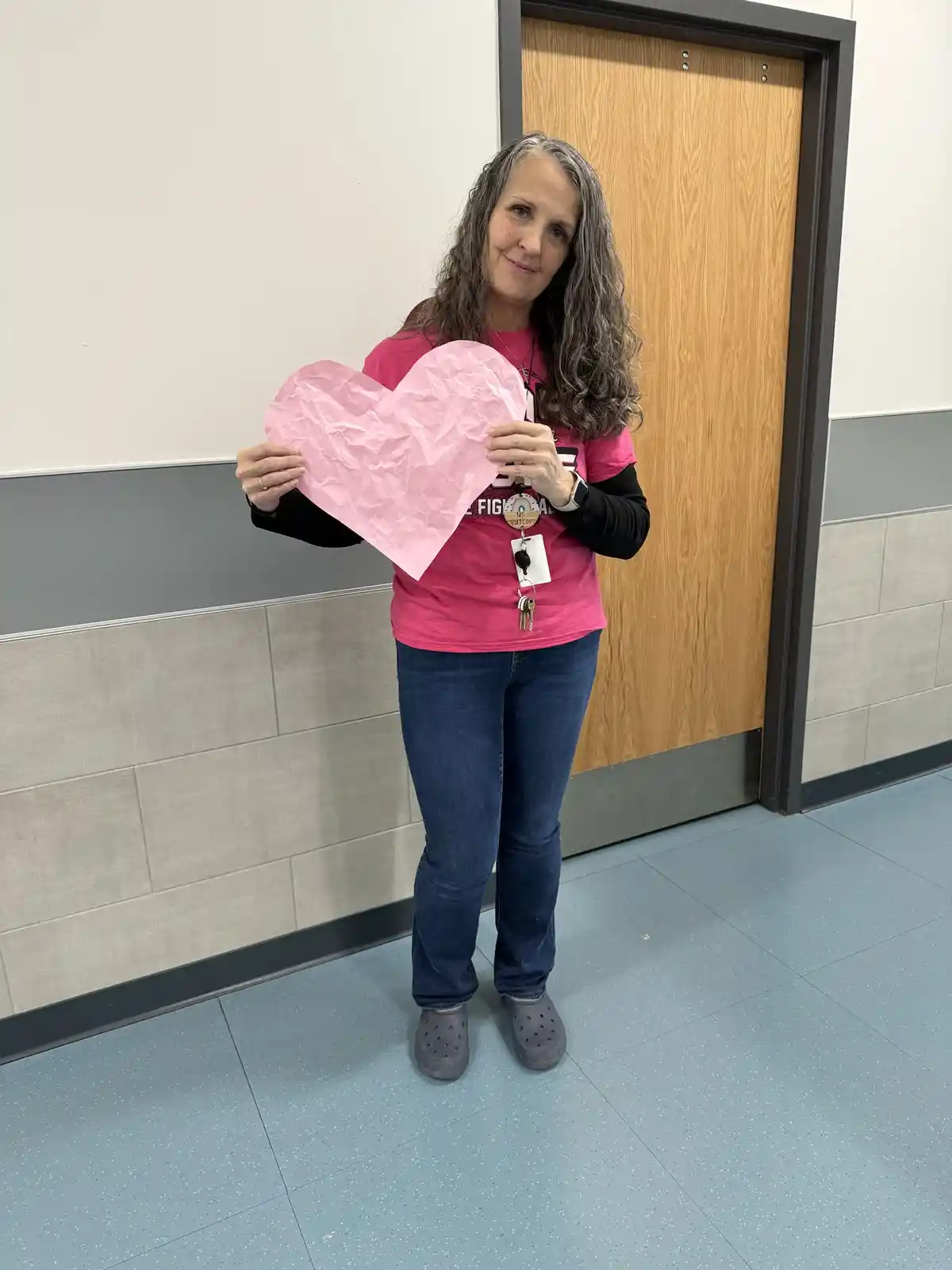 A woman in a pink shirt and jeans stands in a hallway holding a large, crumpled pink paper heart. She has a subtle, knowing…