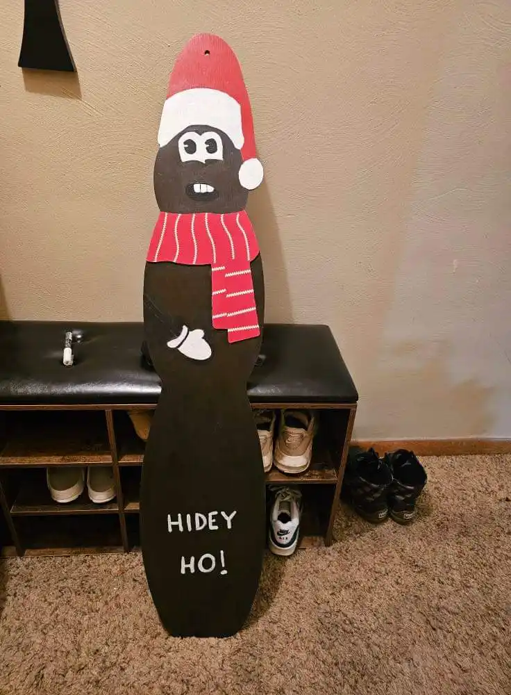 A handmade, dark brown, vertically elongated wooden cutout resembling a person wearing a red and white Santa hat and a red…