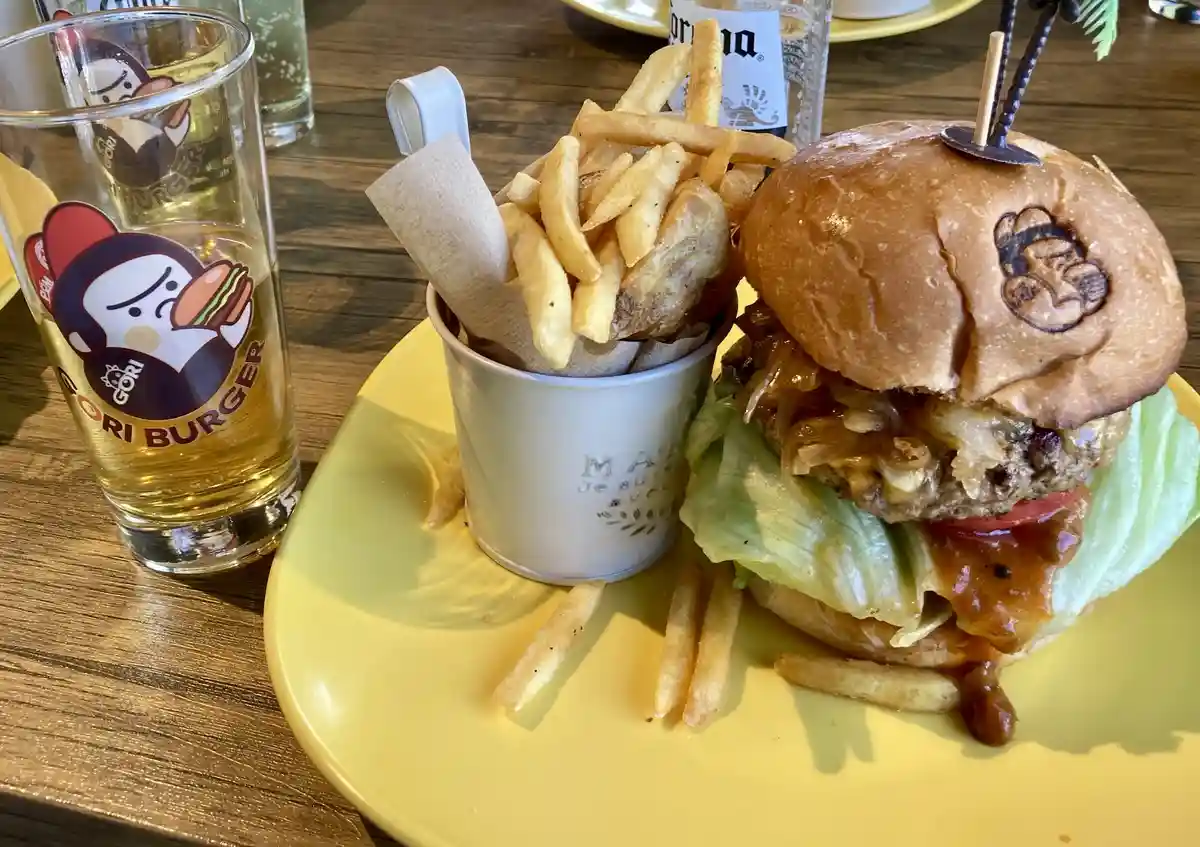 A close-up photo of a large, messy burger with lettuce, tomato, and sauce on a yellow plate, accompanied by a side of fries…