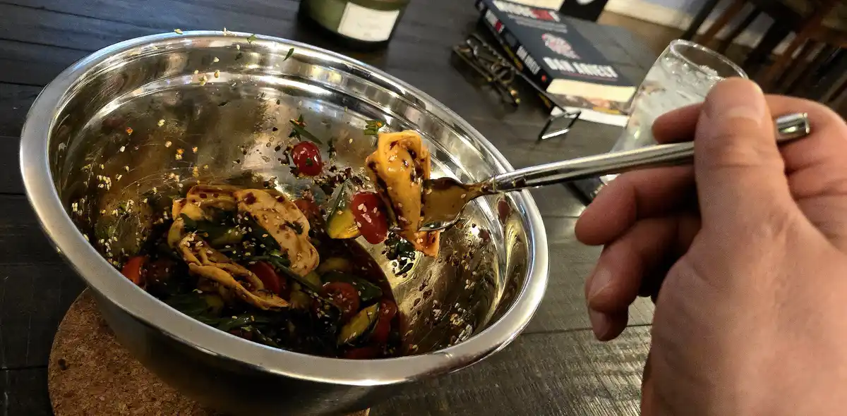 A close-up photo shows a hand holding a spoonful of salad over a nearly empty stainless steel bowl. The salad contains…