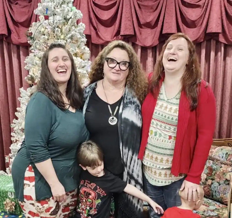 A family photo shows three women and two young boys posing in front of a decorated Christmas tree with red curtains. The…