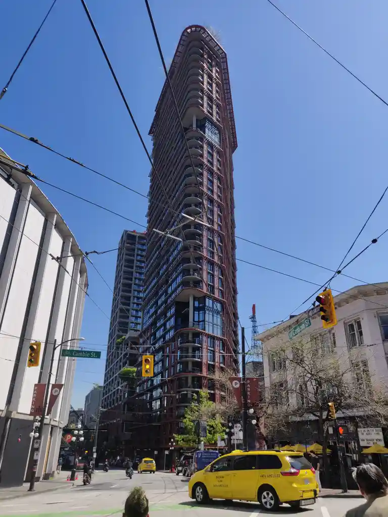 A low-angle street photo shows a tall, uniquely curved modern skyscraper with red and glass facade dominating the view…