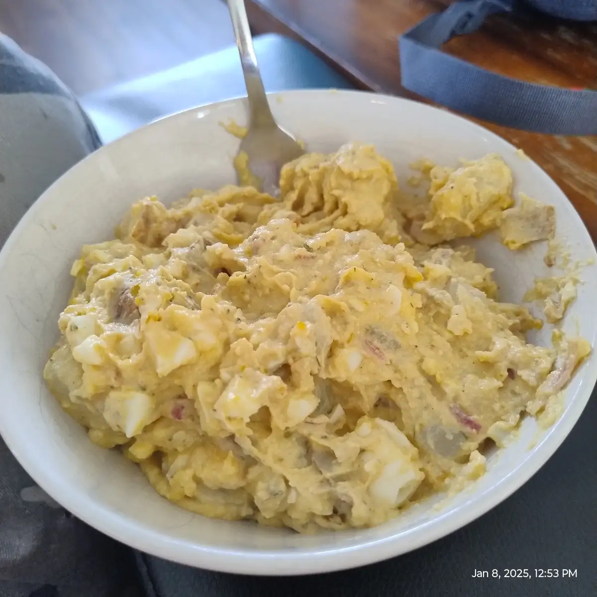 A close-up photo shows a white bowl filled with a creamy, yellowish egg salad, containing visible chunks of hard-boiled egg…