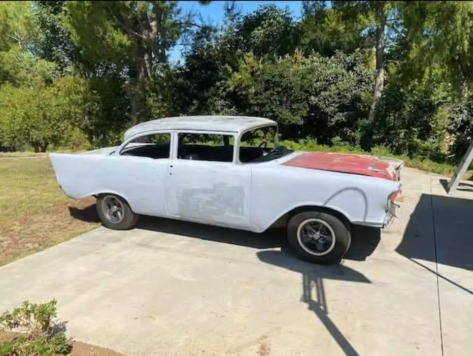 a 1957 chevrolet bel air with a red hood and white body parked on a concrete driveway surrounded by trees and grass. the car…