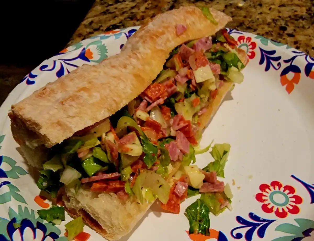 A close-up photo shows a generously filled sandwich on a crusty roll, overflowing with diced ham, salami, lettuce, and other…