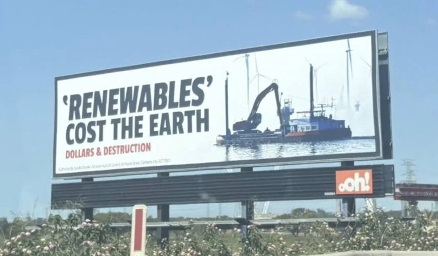 A large outdoor billboard displays the text 'RENEWABLES' COST THE EARTH' in large black letters, with 'DOLLARS &…