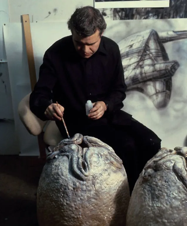 A man in a black shirt sits in an art studio, intently painting a large, textured sculpture of a squid with a brush. Behind…