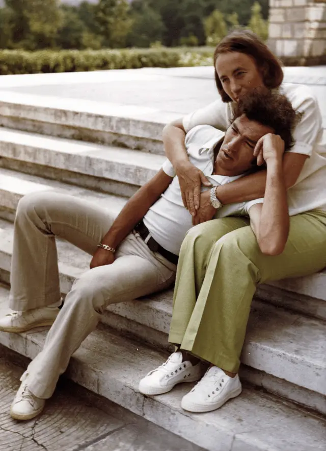 A vintage photo shows a man reclining on stone steps with a woman embracing him from behind; he has his eyes closed and hand…