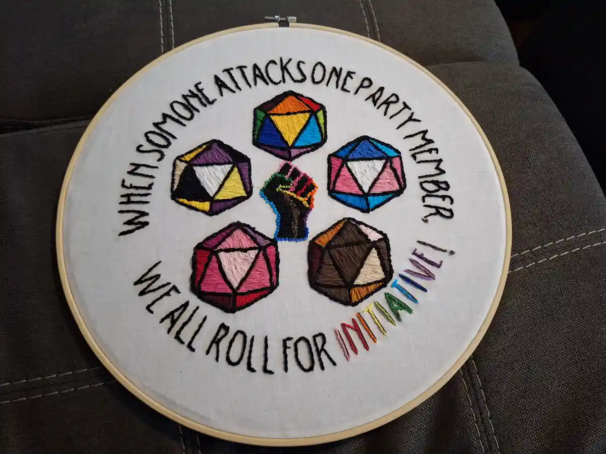 An embroidery hoop displays a hand-stitched design featuring five colorful, multi-sided dice arranged around a central black…