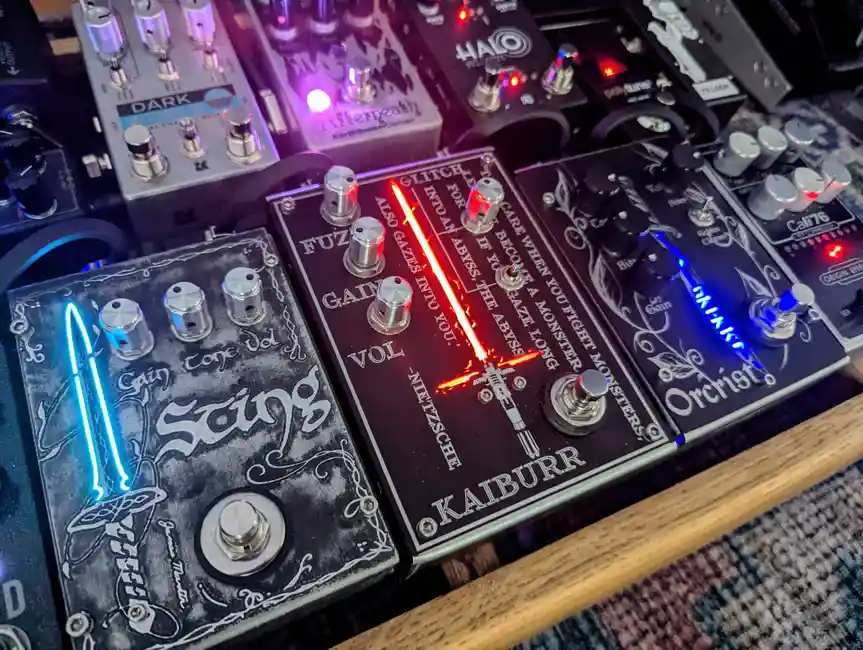 A close-up photo of several guitar effect pedals with glowing LED lights, including a central black pedal labeled 'KAIBURR'…