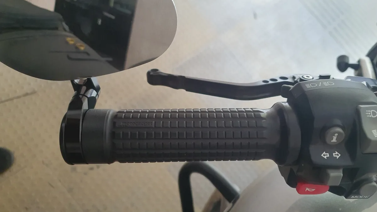 A close-up photo of a motorcycle handlebar with a textured grip and various control buttons, including a red horn button and…