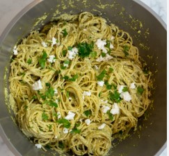 a close-up photo of a metal mixing bowl filled with cooked spaghetti coated in a green sauce, topped with chopped fresh…