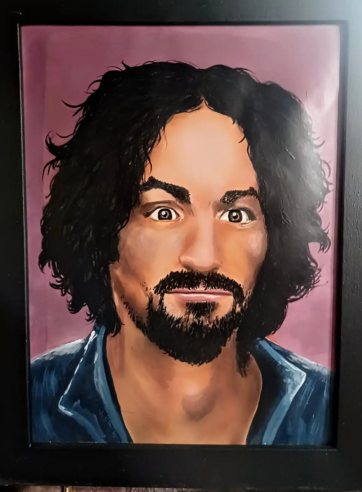 A framed portrait painting depicts a man with long black curly hair, a full beard, and wide, surprised-looking eyes against…