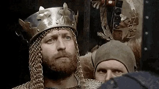 A still from a medieval drama shows a bearded king in a metal crown and chainmail looking sad and pensive. The same image is…