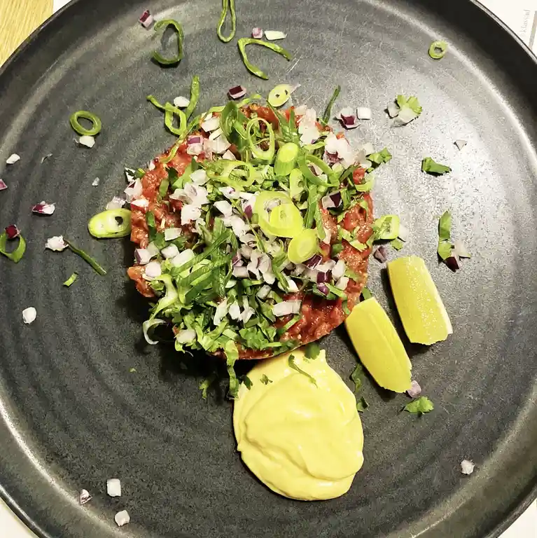 A top-down photo of a dark gray plate holding a raw meat tartare dish, topped with chopped green onions, red onions, and…