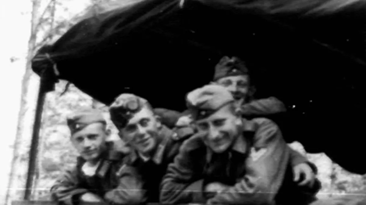 A grainy black-and-white photo shows four smiling men in military uniforms huddled under a tent, with overlaid text reading…