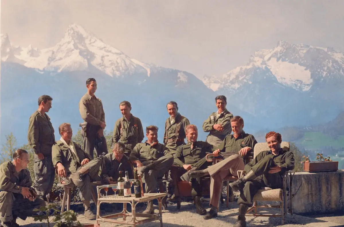 A vintage photograph shows a group of men in military uniforms relaxing outdoors with bottles and glasses on a table, set…