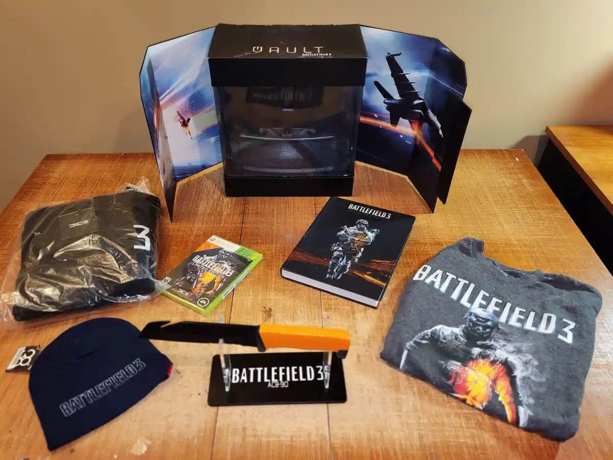 A collection of Battlefield 3 collector's edition merchandise is laid out on a wooden table, including a large 'VAULT'…