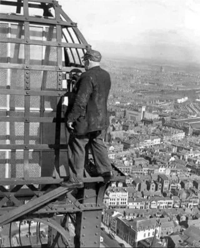 A black-and-white historical photo shows a man in a cap and jacket standing on the exposed metal framework of a tall…