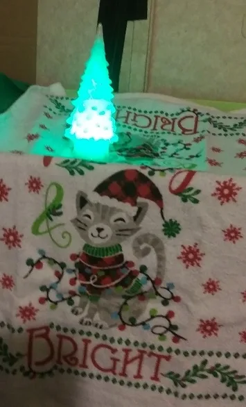 A small, glowing green plastic Christmas tree sits atop a white holiday-themed towel featuring an embroidered cat wearing a…