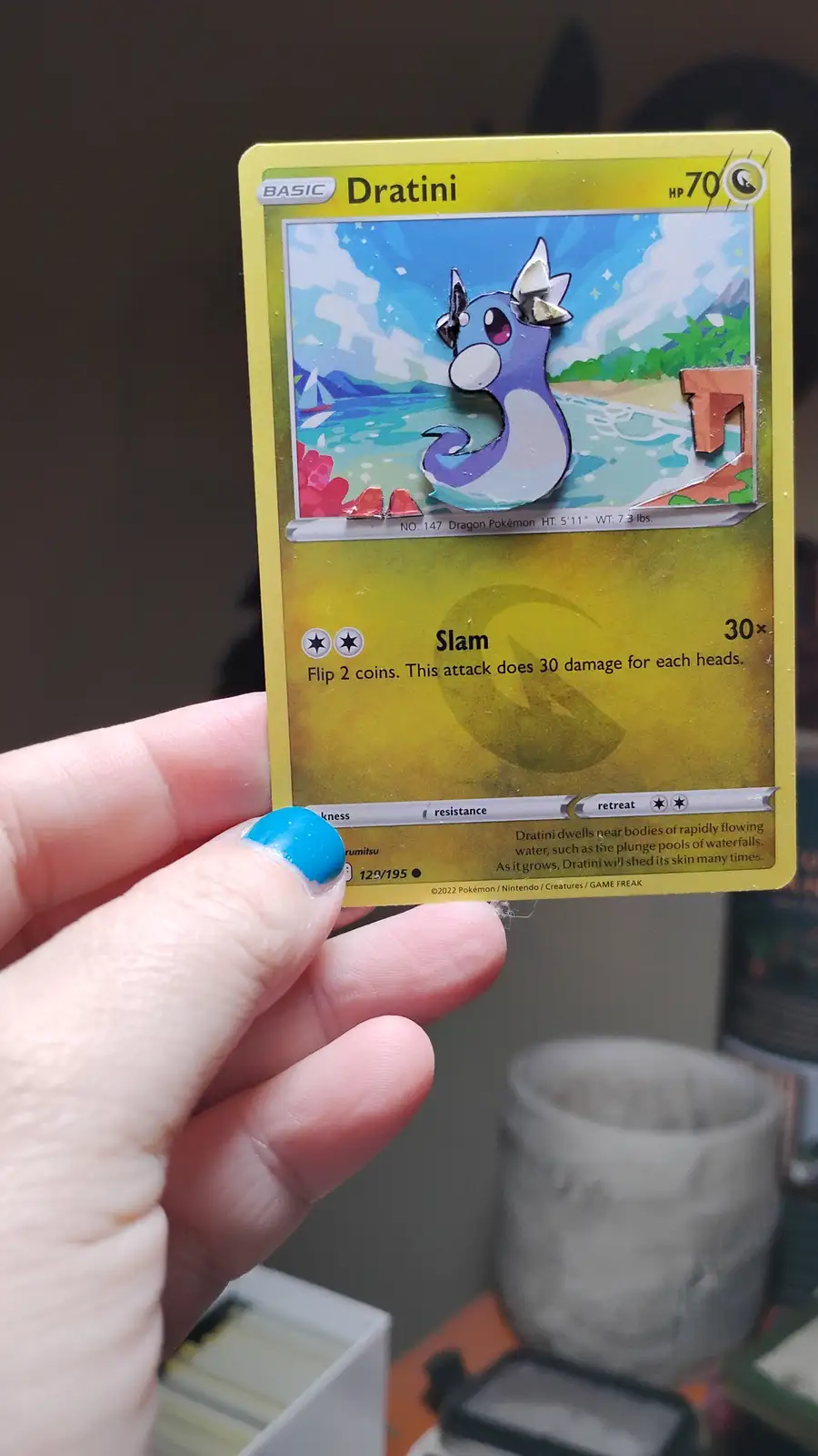 A hand holds a yellow Pokémon card featuring Dratini, which has been physically altered with cutouts and paper pieces to…
