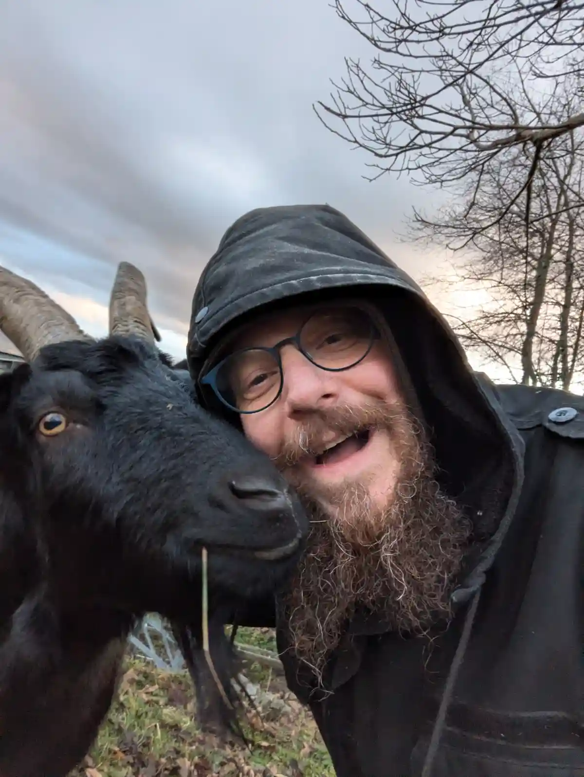 A man with a long beard and glasses, wearing a black hooded jacket, takes a selfie with a black goat that has large curved…