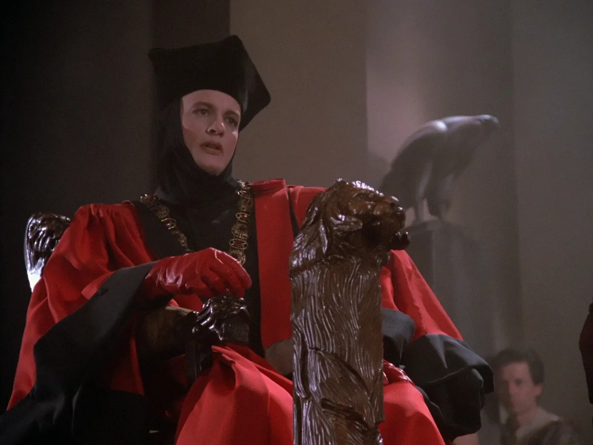 a man in a red and black robe with a black hood and red gloves sits on a throne with lion-head armrests, looking upward with…