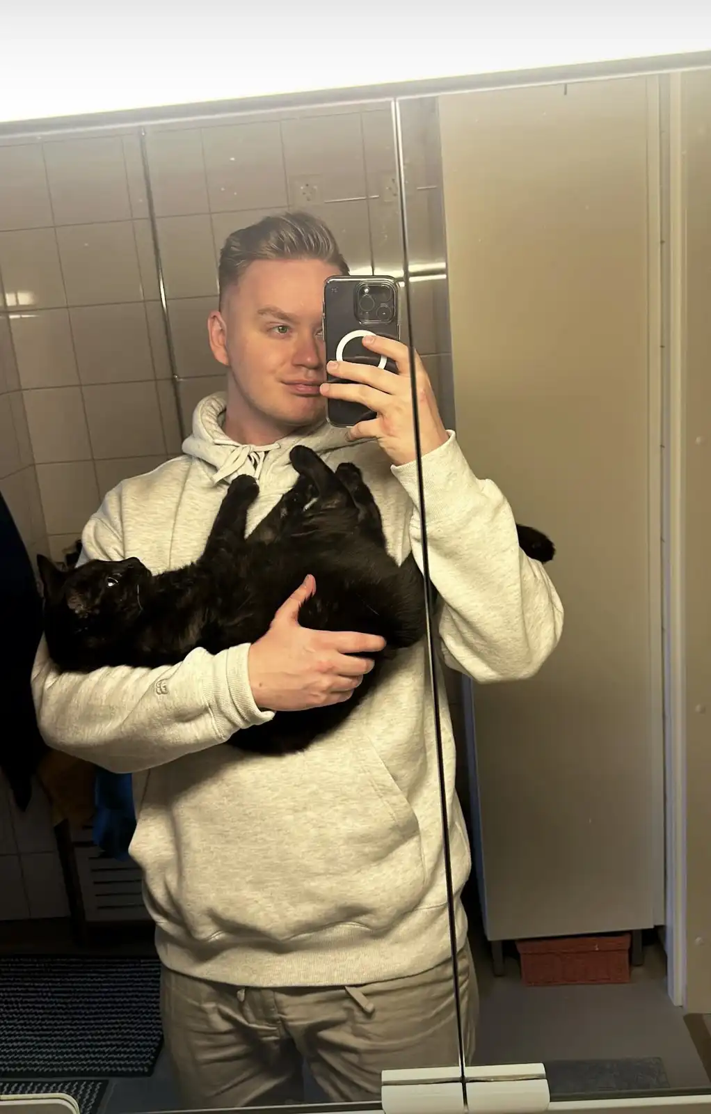 A young man in a light grey hoodie takes a mirror selfie in a bathroom while holding a black cat that is lying on its back…