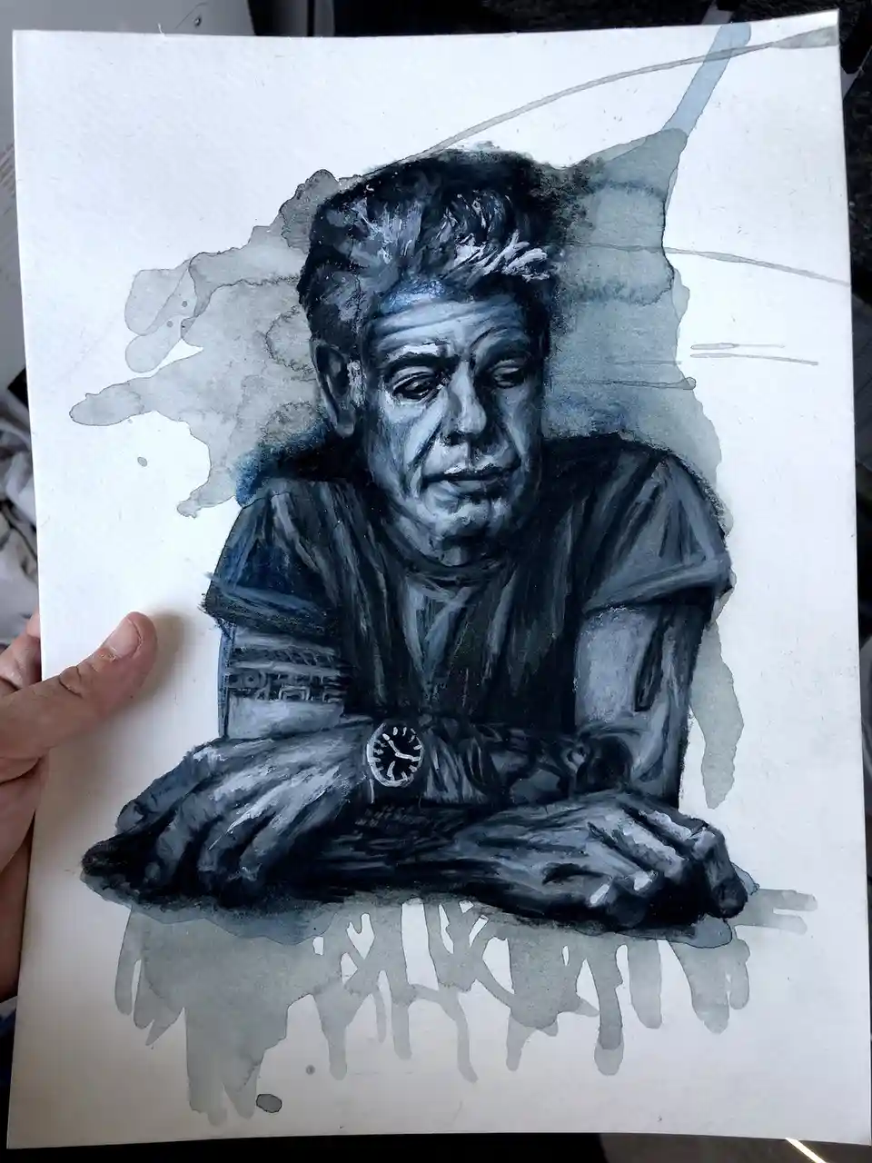 A hand holds a watercolor portrait of a man with a weary expression, arms crossed on a surface, wearing a watch. The artwork…