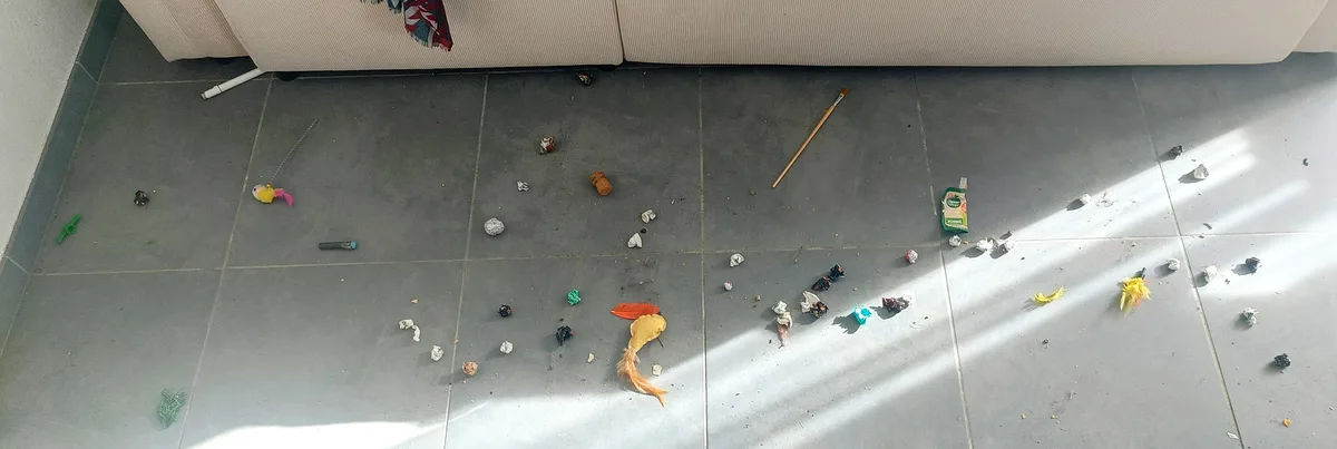 A wide-angle shot of a grey tiled floor littered with cat toys, crumpled paper, and scattered debris, with a beige sofa in…