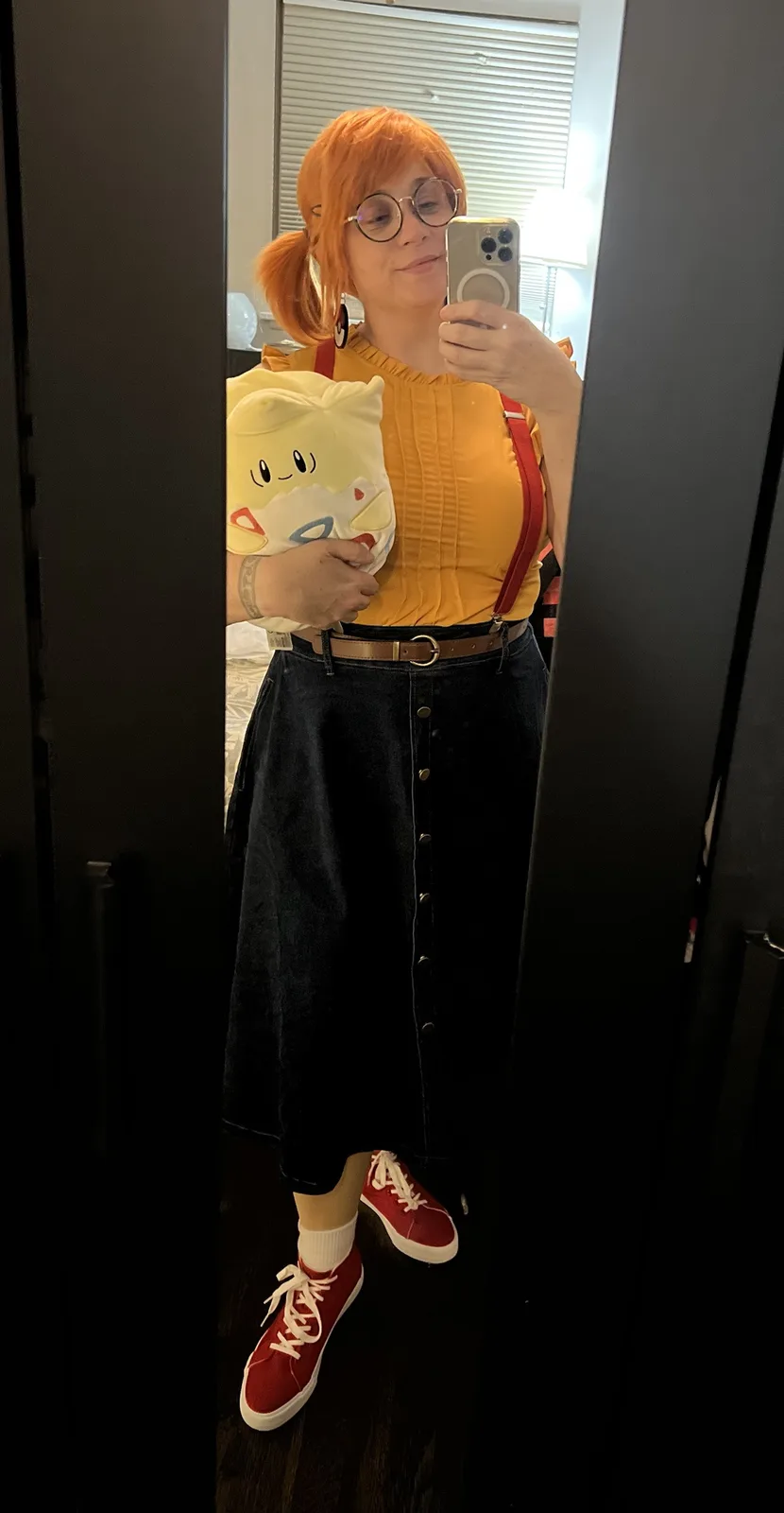 A person with orange pigtails and glasses takes a mirror selfie while holding a plush of the Pokémon Pikachu. They wear a…