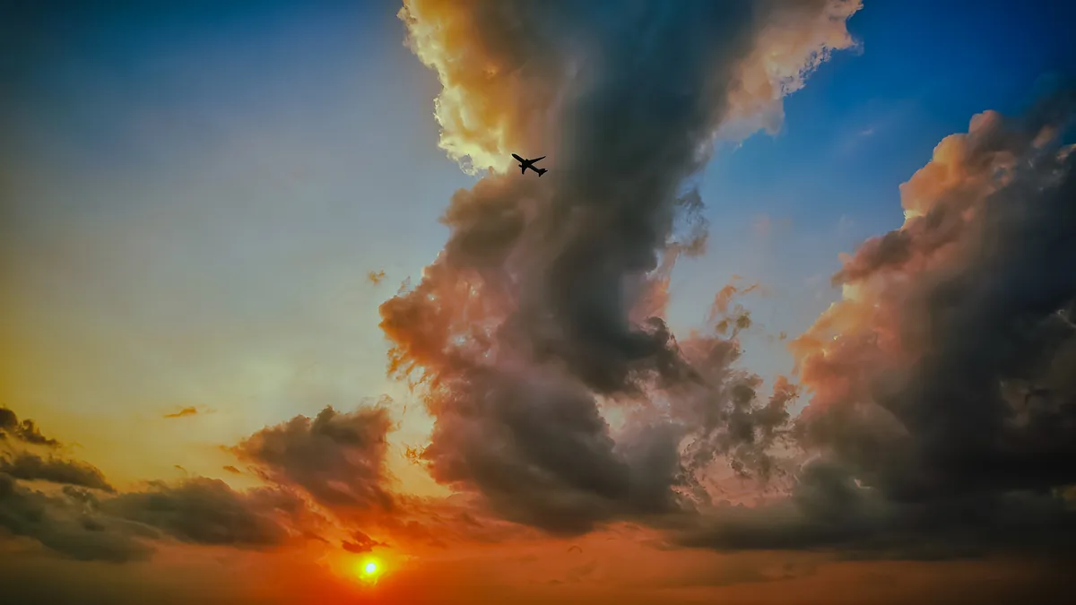 A dramatic sunset sky with orange and blue hues, featuring large, textured clouds and a bright sun near the horizon. A small…