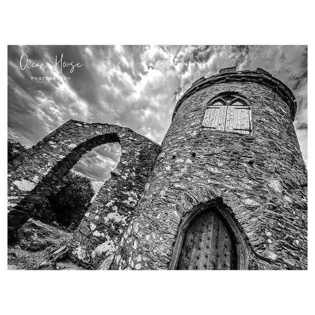 A black-and-white photograph shows a low-angle view of a stone tower with a wooden door and a boarded-up arched window, next…