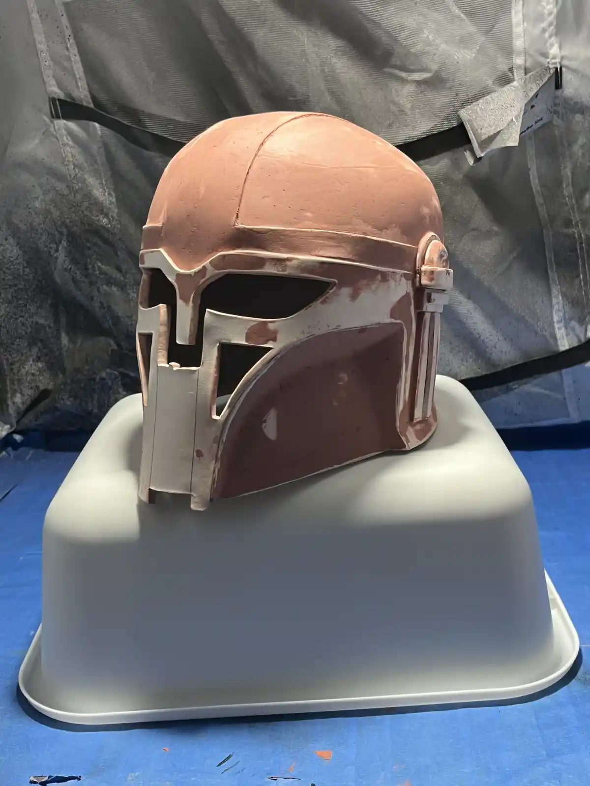 A Mandalorian helmet, likely a cosplay prop, is shown in an unfinished state, resting on a white plastic bowl. The helmet is…