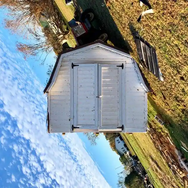 A tilted photo of a white wooden shed with double doors, set in a grassy yard with scattered debris and a red tractor in the…