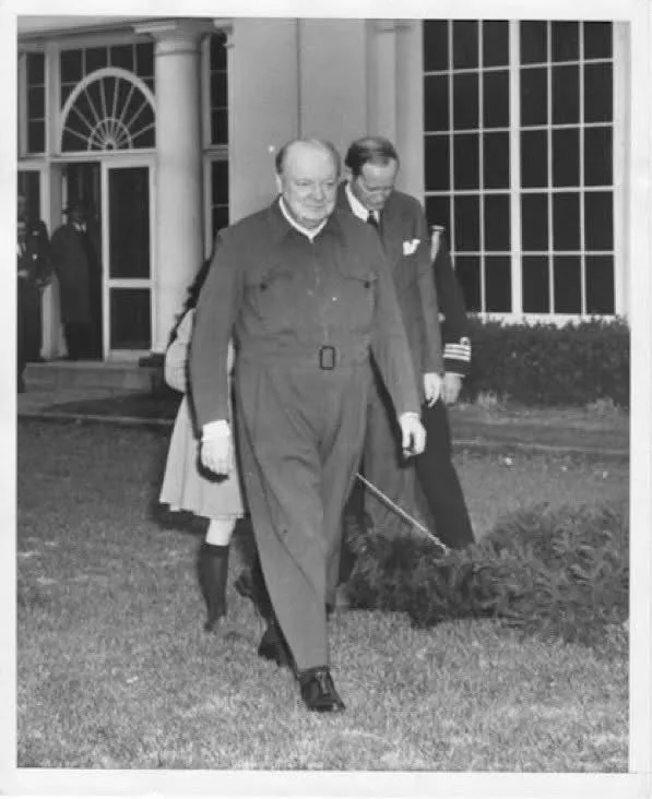 A black-and-white photograph shows Winston Churchill walking outdoors on grass, holding a cane in his right hand. He is…
