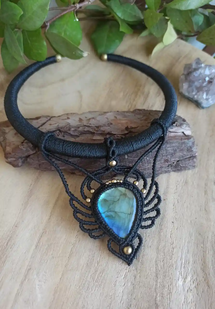 A black macrame necklace with a teardrop-shaped labradorite stone pendant and gold beads rests on a piece of driftwood on a…