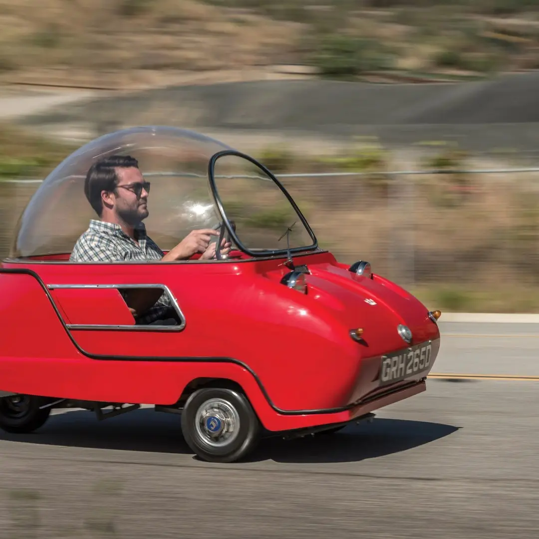 A man with a beard and sunglasses drives a red, bubble-topped, three-wheeled vehicle with the license plate 'GRH 2650' on a…
