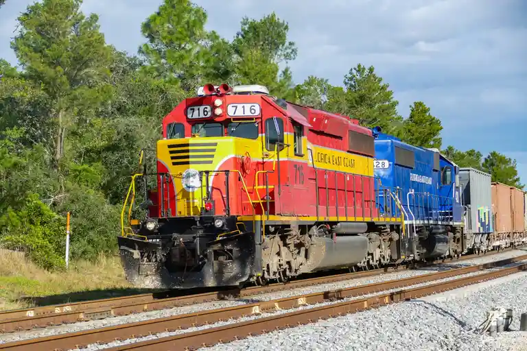 A red and yellow Florida East Coast locomotive numbered 716 is coupled to a blue locomotive numbered 28, both pulling…