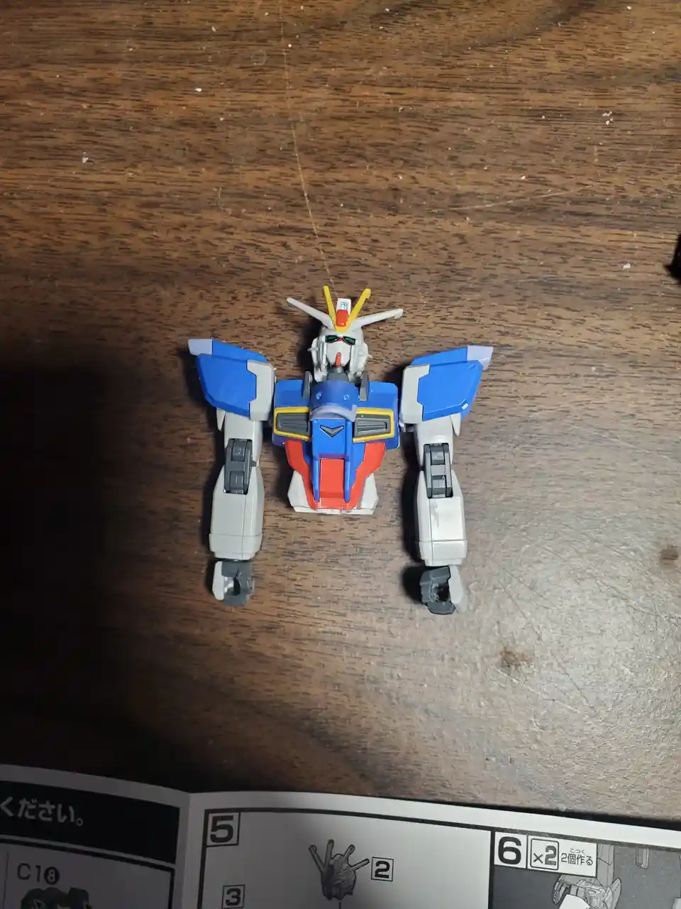 A small blue, white, and red Gundam-style robot toy sits on a wooden table, viewed from above, with an open instruction…