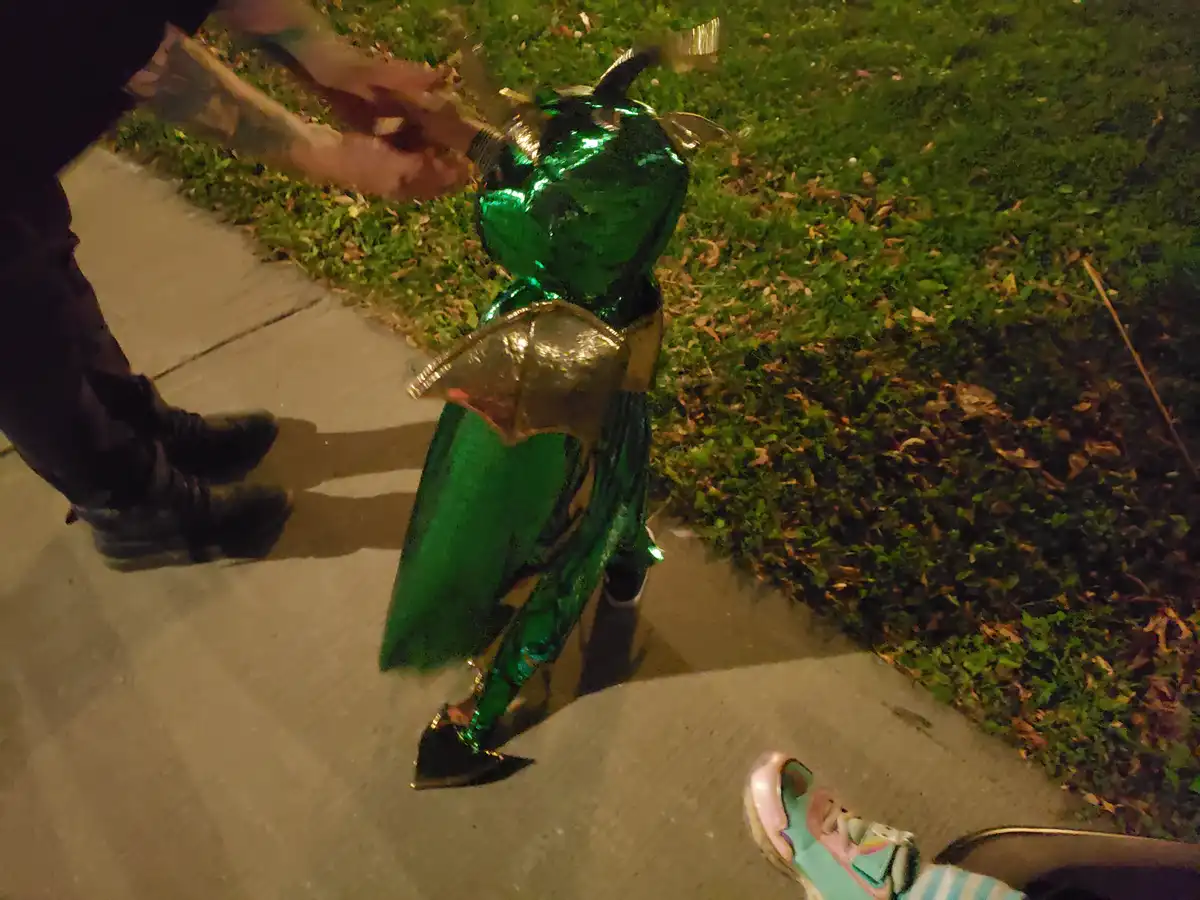 A child in a shiny green dragon costume with gold wings and horns stands on a sidewalk at night, while an adult with tattoos…