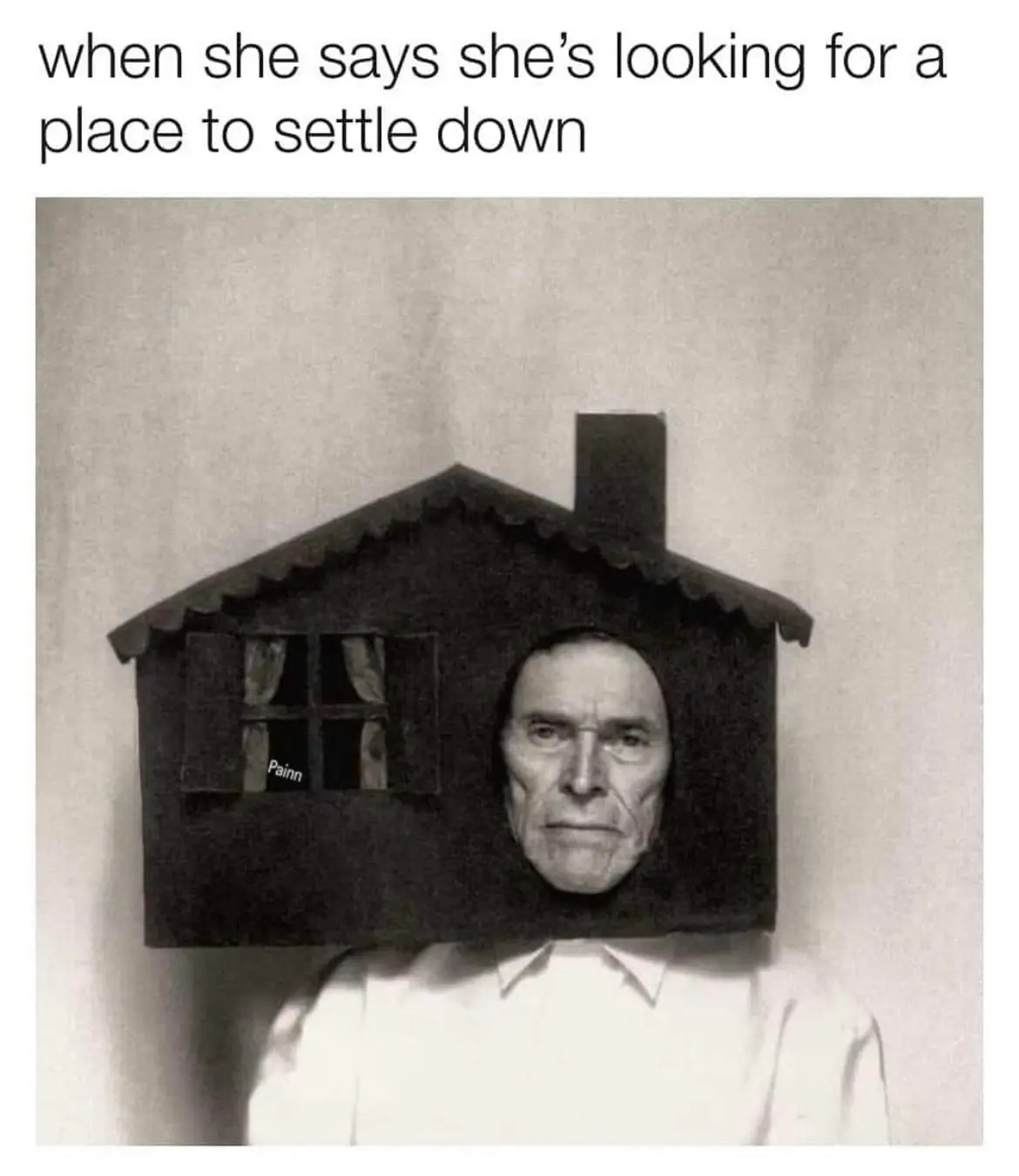 A black-and-white photo shows a man wearing a small, dark house as a headpiece, with a window on the side and a chimney on…