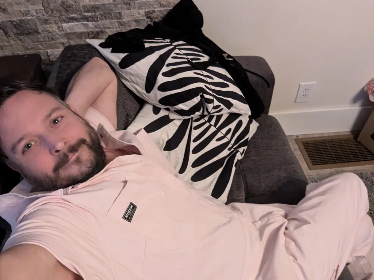 A man with a beard wearing a light pink shirt and matching pants lies on a gray couch, propped up by black-and-white…