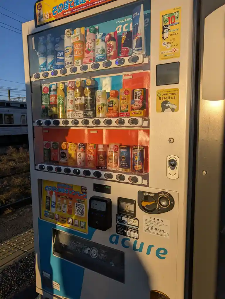 A Japanese vending machine branded 'acure' is shown at a train station platform, filled with various drinks like Red Bull…