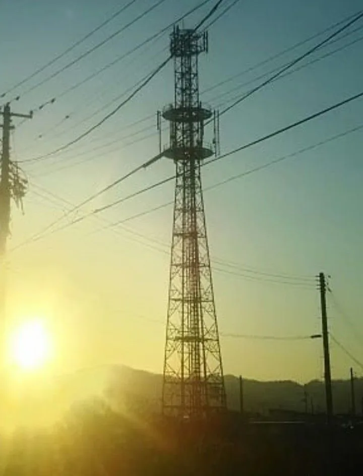 A tall, metal telecommunications tower stands silhouetted against a bright, low-hanging sun during sunrise or sunset, with…